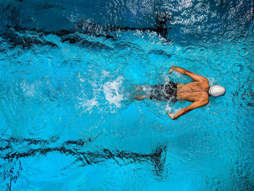 Top view of a swimmer wearing a cap, performing a front crawl stroke in a clear blue swimming pool.